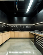 A clean industrial garage featuring polished concrete floors, birch plywood cabinets, and black perforated steel wall panels for tool organization. Overhead LED strip lights illuminate a stainless steel workbench, with a few carefully arranged silver and black tools. — styling and décor detail