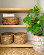 Cozy boho laundry room featuring light oak floating shelves, natural rattan laundry baskets, a woven jute rug, and a ceramic planter with a lush Pothos plant, all under soft, warm lighting. — alternate angle