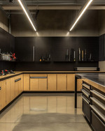 A clean industrial garage featuring polished concrete floors, birch plywood cabinets, and black perforated steel wall panels for tool organization. Overhead LED strip lights illuminate a stainless steel workbench, with a few carefully arranged silver and black tools. — close-up of textures and materials