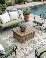 A boho poolside area featuring a mix of vintage wrought-iron chairs in sage green and a modern low-slung outdoor sofa with natural linen cushions. A reclaimed wooden coffee table sits on a layered outdoor rug with geometric patterns, surrounded by potted succulents and woven rattan accents. — styling and décor detail