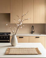 A serene Japandi kitchen featuring light ash wood cabinetry, a white quartz countertop, and minimalist black metal fixtures. A single ceramic vase holds delicate cherry blossom branches, and a natural woven placemat sits on the island. — close-up of textures and materials