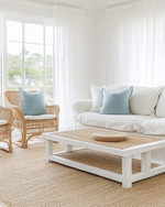 A serene coastal sunroom featuring a slipcovered sofa in a creamy off-white, paired with rattan chairs and a bleached oak coffee table. Soft dusty blue throw pillows and a woven jute rug add subtle color accents, all illuminated by ample natural light filtering through sheer linen curtains. — close-up of textures and materials