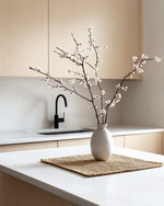 A serene Japandi kitchen featuring light ash wood cabinetry, a white quartz countertop, and minimalist black metal fixtures. A single ceramic vase holds delicate cherry blossom branches, and a natural woven placemat sits on the island. — alternate angle