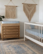Coastal baby and kids' room with a white oak crib, a large seagrass rug, a rattan changing table, and a macrame wall hanging above a dresser. Soft white walls are accented with muted blue textiles and natural wood elements. — close-up of textures and materials