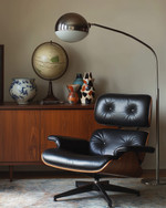 A sophisticated Mid-Century Modern library featuring an Eames Lounge Chair in black leather and walnut, an Arco floor lamp arching over, and a teak credenza displaying abstract ceramic vases and a vintage globe, all under the warm glow of a Sputnik chandelier.
