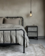 An industrial bedroom featuring a black metal bed frame, a grey washed linen duvet, and an exposed Edison bulb pendant light hanging from the ceiling. A dark wood nightstand holds a concrete-look lamp, and a large, distressed area rug covers the light grey floor. — styling and décor detail