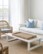 A serene coastal sunroom featuring a slipcovered sofa in a creamy off-white, paired with rattan chairs and a bleached oak coffee table. Soft dusty blue throw pillows and a woven jute rug add subtle color accents, all illuminated by ample natural light filtering through sheer linen curtains. — styling and décor detail