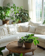 A cozy boho family room featuring a cream sectional sofa adorned with macrame and striped throw pillows, a round rattan coffee table, and an abundance of lush green plants in woven and ceramic planters, all bathed in natural light from a large window with sheer linen curtains. — alternate angle