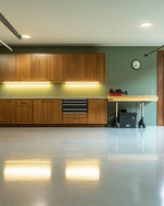 Clean Mid-Century Modern garage featuring walnut flat-panel cabinetry, a polished light grey epoxy floor, recessed LED lighting, and a subtle sage green accent wall behind a minimalist workbench. — alternate angle