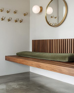 A Mid-Century Modern mudroom featuring a walnut slatted bench, polished concrete flooring, and an olive green upholstered cushion. Brass hooks line the wall above, with a circular mirror and a small white globe sconce adding modern accents.