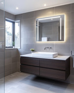 Modern bathroom with a floating dark wood vanity, white vessel sink, and large format grey wall tiles. Integrated LED mirror lighting and recessed ceiling lights illuminate the space, complemented by polished chrome fixtures. — alternate angle