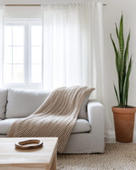 A bright Scandinavian living room with white walls, a light grey sofa, a simple birch wood coffee table, and a chunky knit throw in oatmeal. Sheer white linen curtains frame a window, and a tall Snake Plant in a terracotta pot adds greenery. — close-up of textures and materials