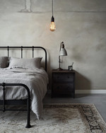 An industrial bedroom featuring a black metal bed frame, a grey washed linen duvet, and an exposed Edison bulb pendant light hanging from the ceiling. A dark wood nightstand holds a concrete-look lamp, and a large, distressed area rug covers the light grey floor. — alternate angle