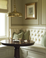 A cozy traditional breakfast nook featuring a built-in cream banquette with sage green cushions, a round dark wood pedestal table, and a classic brass pendant light, all bathed in soft morning light. — alternate angle
