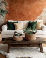 A cozy rustic family room featuring a warm terracotta accent wall behind a cream linen sofa adorned with forest green and oatmeal throw pillows. A distressed wooden coffee table sits on a faux sheepskin rug, while woven baskets and ceramic planters with greenery add natural elements.