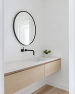 A bright Scandinavian powder room with white walls, a light wood floating vanity, a minimalist round mirror, and a single black wall-mounted faucet, accented by a small green plant. — alternate angle