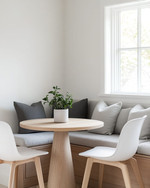 A small, bright Scandinavian breakfast nook featuring a light wood pedestal table, two minimalist white chairs, and a built-in banquette with grey linen cushions. A small potted plant sits on the table, and white walls create an airy, open feel. — close-up of textures and materials