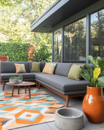 A family-friendly Mid-Century Modern patio featuring a low-profile charcoal grey L-shaped sectional with olive green performance fabric cushions, a geometric outdoor rug in burnt orange and teal, and teak wood accent tables. Large, sculptural ceramic planters with pet-safe plants are arranged around the seating area, with a subtle concrete water bowl for pets integrated into the design. — styling and décor detail