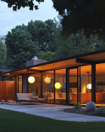 A Mid-Century Modern exterior at dusk, featuring a low-slung roofline, large glass windows, and warm glow from spherical globe sconces flanking the entry. A Sputnik chandelier is visible under a covered patio, illuminating a seating area with clean-lined furniture and a natural wood fence. — alternate angle