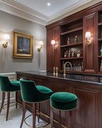 A traditional home bar featuring rich cherry wood cabinetry, a polished brass bar faucet, and a black marble countertop. Walls are painted in a soft greige, and the space is illuminated by classic wall sconces and recessed lighting, with emerald green velvet bar stools. — styling and décor detail