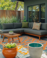 A family-friendly Mid-Century Modern patio featuring a low-profile charcoal grey L-shaped sectional with olive green performance fabric cushions, a geometric outdoor rug in burnt orange and teal, and teak wood accent tables. Large, sculptural ceramic planters with pet-safe plants are arranged around the seating area, with a subtle concrete water bowl for pets integrated into the design.