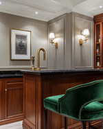 A traditional home bar featuring rich cherry wood cabinetry, a polished brass bar faucet, and a black marble countertop. Walls are painted in a soft greige, and the space is illuminated by classic wall sconces and recessed lighting, with emerald green velvet bar stools. — alternate angle