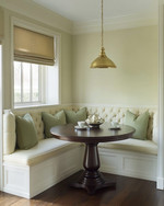 A cozy traditional breakfast nook featuring a built-in cream banquette with sage green cushions, a round dark wood pedestal table, and a classic brass pendant light, all bathed in soft morning light. — styling and décor detail