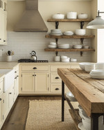 A cozy rustic kitchen featuring creamy off-white cabinets, a reclaimed wood island, and open shelving displaying white stoneware. Wrought iron hardware and a sisal runner add to the warm, inviting atmosphere, with natural light streaming in.