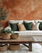A cozy rustic family room featuring a warm terracotta accent wall behind a cream linen sofa adorned with forest green and oatmeal throw pillows. A distressed wooden coffee table sits on a faux sheepskin rug, while woven baskets and ceramic planters with greenery add natural elements. — alternate angle