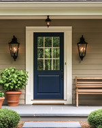 A cozy traditional exterior featuring a warm gray facade with creamy white trim, a deep blue front door, and classic black lantern sconces flanking the entrance. Potted green plants in terracotta pots sit on either side of the door, and a traditional wooden bench offers a welcoming touch.