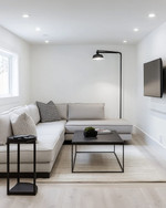 A small modern basement living area featuring a light gray modular sectional sofa, a black metal coffee table, and a wall-mounted television. White walls and light wood flooring contribute to an open, airy feel, complemented by recessed lighting and a tall, slender floor lamp. — alternate angle