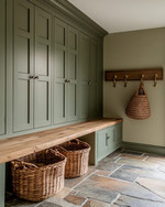 A traditional mudroom with sage green built-in cabinetry, a natural wood bench, and flagstone flooring. Woven rattan baskets are neatly tucked under the bench, and aged brass hooks line the wall.