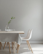 A bright Scandinavian dining room featuring a light birch wood dining table, minimalist white chairs, a simple linen tablecloth, and a clear glass vase with a single green branch, all set against soft grey walls.