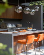 A stylish Mid-Century Modern outdoor kitchen featuring a grey terrazzo countertop, rich walnut-toned cabinetry, and a sleek brushed stainless steel grill. Orange upholstered bar stools with chrome legs sit at the island, under a cluster of spherical globe pendant lights. — styling and décor detail