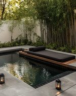A serene Japandi pool area featuring a dark-tiled swimming pool surrounded by smooth concrete pavers and a low-slung teak daybed with charcoal cushions. Subtle solar-powered lanterns glow amidst pet-friendly green groundcover and slender bamboo plants. — styling and décor detail