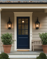 A cozy traditional exterior featuring a warm gray facade with creamy white trim, a deep blue front door, and classic black lantern sconces flanking the entrance. Potted green plants in terracotta pots sit on either side of the door, and a traditional wooden bench offers a welcoming touch. — alternate angle