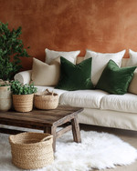 A cozy rustic family room featuring a warm terracotta accent wall behind a cream linen sofa adorned with forest green and oatmeal throw pillows. A distressed wooden coffee table sits on a faux sheepskin rug, while woven baskets and ceramic planters with greenery add natural elements. — styling and décor detail