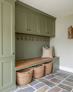 A traditional mudroom with sage green built-in cabinetry, a natural wood bench, and flagstone flooring. Woven rattan baskets are neatly tucked under the bench, and aged brass hooks line the wall. — styling and décor detail