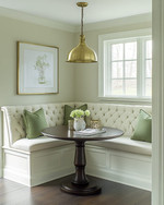 A cozy traditional breakfast nook featuring a built-in cream banquette with sage green cushions, a round dark wood pedestal table, and a classic brass pendant light, all bathed in soft morning light.