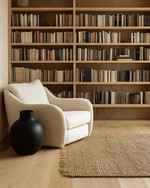 A Japandi library featuring light blonde wood bookshelves filled with books, a comfortable cream linen armchair, and a natural jute rug on light hardwood floors, accented by a matte black ceramic vase. — alternate angle