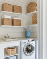 Small coastal laundry room featuring white shiplap walls, a stackable washer and dryer, open shelving with woven baskets, and a light quartz countertop with a small farmhouse sink. Soft blue accents are visible in a decorative ceramic vase. — alternate angle