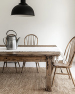 A bright farmhouse dining room featuring a distressed oak dining table, spindle-back chairs, and a woven rug. A large, vintage-inspired matte black pendant light hangs above, with a galvanized metal pitcher and white linen runner on the table. Walls are painted in a creamy off-white. — close-up of textures and materials
