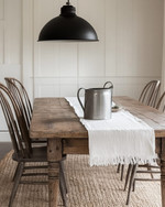 A bright farmhouse dining room featuring a distressed oak dining table, spindle-back chairs, and a woven rug. A large, vintage-inspired matte black pendant light hangs above, with a galvanized metal pitcher and white linen runner on the table. Walls are painted in a creamy off-white. — alternate angle