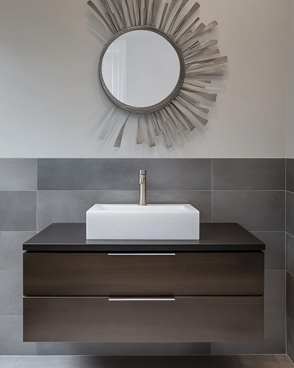 A modern powder room features a floating dark wood vanity with an integrated white quartz sink and brushed nickel faucet, set against large format charcoal grey porcelain tiles. A simple round mirror and abstract metal wall art complete the sleek, resilient design.