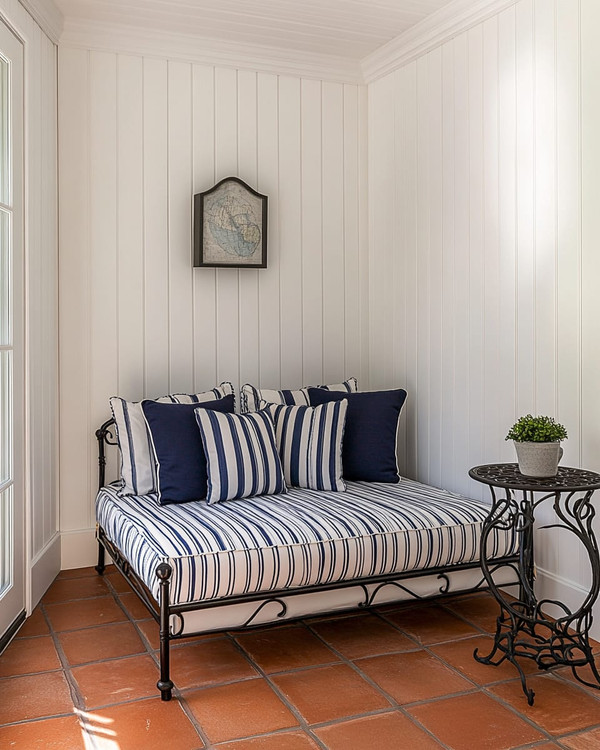 A compact traditional poolhouse interior featuring white beadboard walls, a small daybed with navy and white striped cushions, a wrought iron side table, and terracotta tile flooring, bathed in natural light.