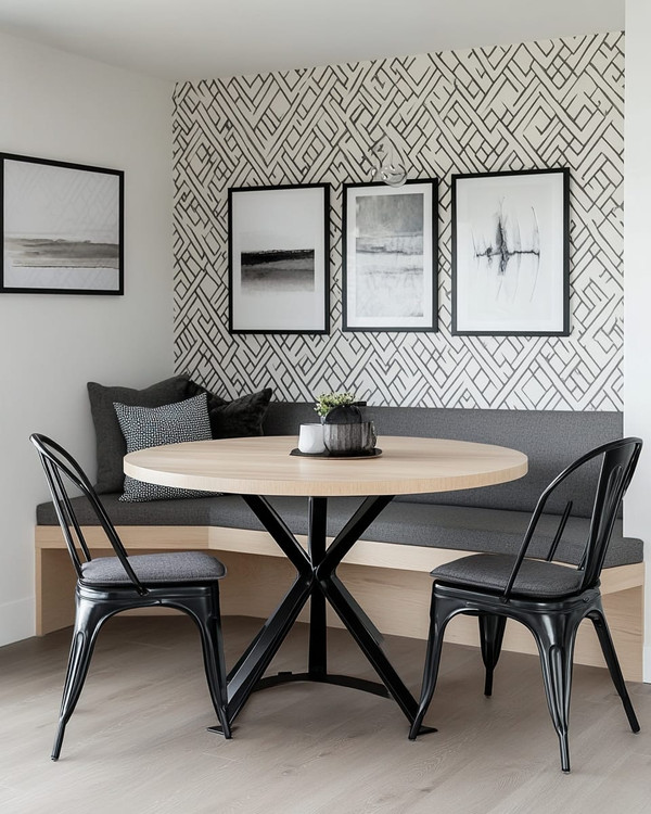 A modern breakfast nook features a light wood round table with a minimalist black metal base, paired with a charcoal grey upholstered bench and two black metal chairs. A subtle geometric peel-and-stick wallpaper adorns the wall, alongside a gallery of abstract art prints.