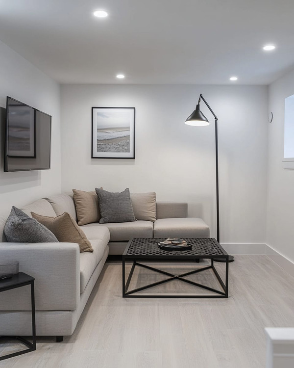 A small modern basement living area featuring a light gray modular sectional sofa, a black metal coffee table, and a wall-mounted television. White walls and light wood flooring contribute to an open, airy feel, complemented by recessed lighting and a tall, slender floor lamp.
