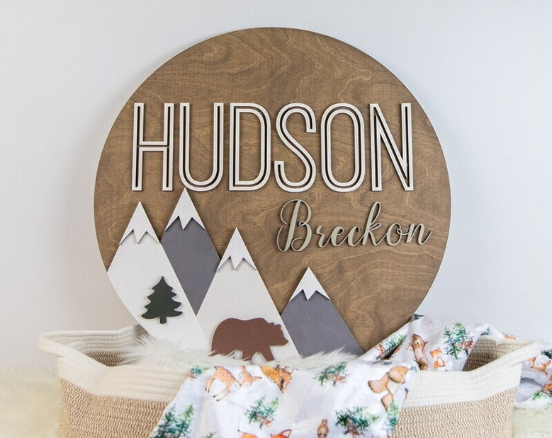 3D mountain woodland round wood nursery sign with personalized baby name, layered trees and peaks, oak finish for nursery wall