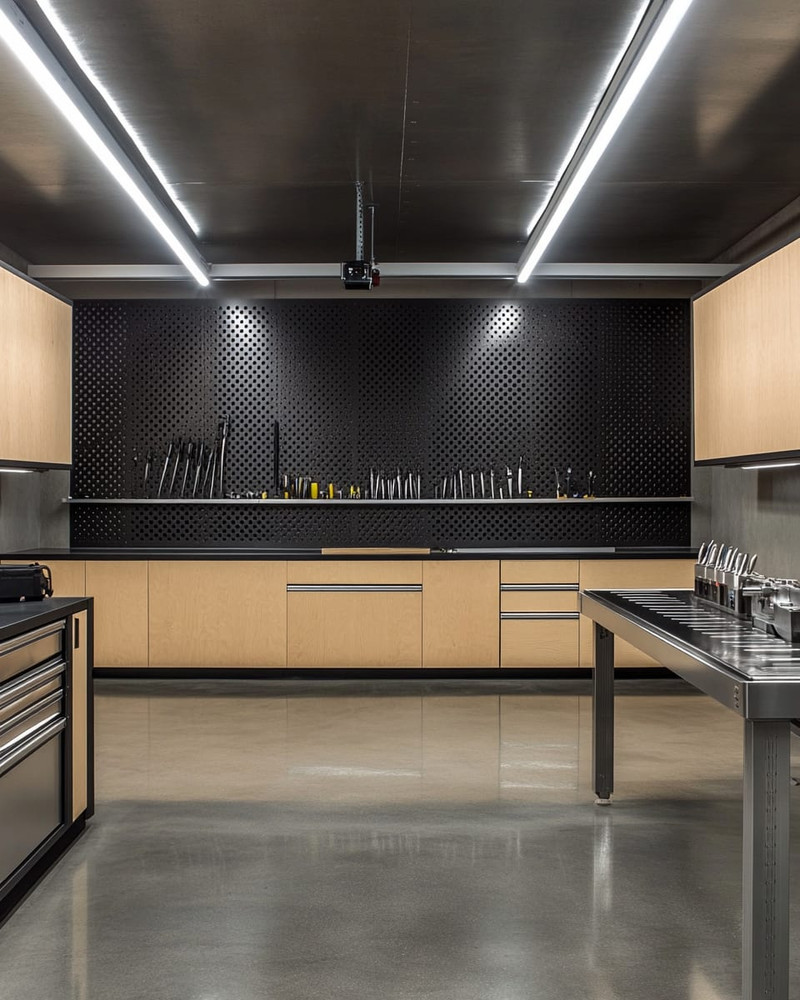 A clean industrial garage featuring polished concrete floors, birch plywood cabinets, and black perforated steel wall panels for tool organization. Overhead LED strip lights illuminate a stainless steel workbench, with a few carefully arranged silver and black tools. A clean industrial garage featuring polished concrete floors, birch plywood cabinets, and black perforated steel wall panels for tool organization. Overhead LED strip lights illuminate a stainless steel workbench, with a few carefully arranged silver and black tools.